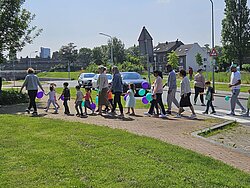 Een groep kinderen en volwassenen die aan het wandelen zijn met ballonnen over een stoep voor een grasveldje